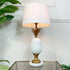 A vintage table lamp shaped like a pineapple, with a ceramic white body, a gold-toned stem and base, and a white drum shade, shown lit on a glass tabletop against a white wall with green plants in the background.