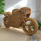 A large decorative rattan motorcycle , photographed indoors on a light rug against a white wall with plants nearby.