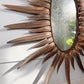 A decorative sunburst wall mirror with a metallic bronze-finished frame resembling stylized feathers or rays, shown mounted on a white paneled wall.