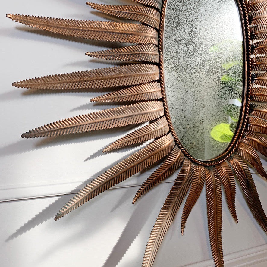A decorative sunburst wall mirror with a metallic bronze-finished frame resembling stylized feathers or rays, shown mounted on a white paneled wall.
