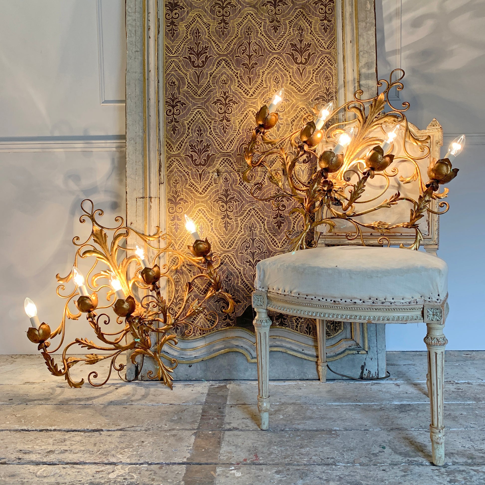 Two ornate gold-toned multi-arm candelabra-style wall lights with lit candle bulbs, displayed in a vintage-style room beside and in front of an upholstered chair and patterned wallpaper.