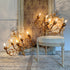 Two ornate gold-toned multi-arm candelabra-style wall lights with lit candle bulbs, displayed in a vintage-style room beside and in front of an upholstered chair and patterned wallpaper.