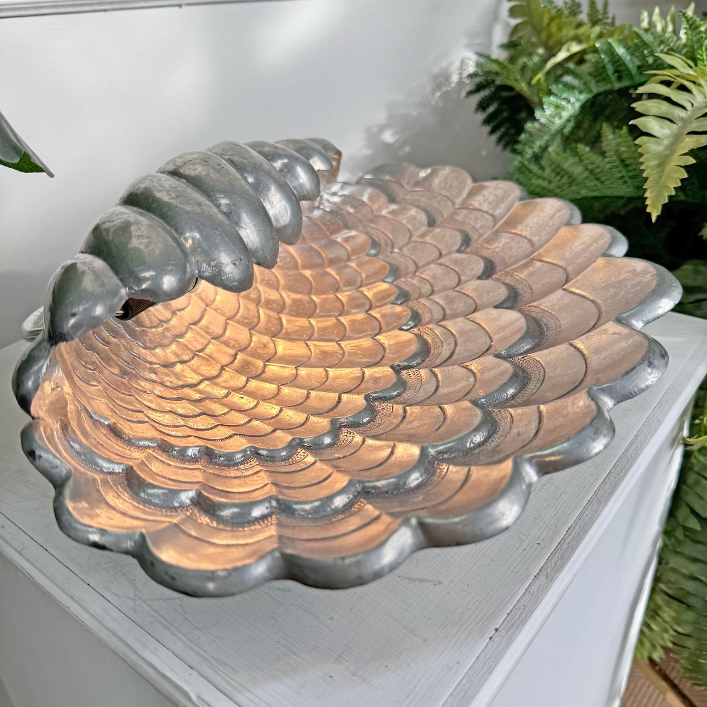 A Vincent Garnier scalloped metal shell lamp with a textured, scale-like pattern, shown resting on a white wooden surface with green plants in the background.