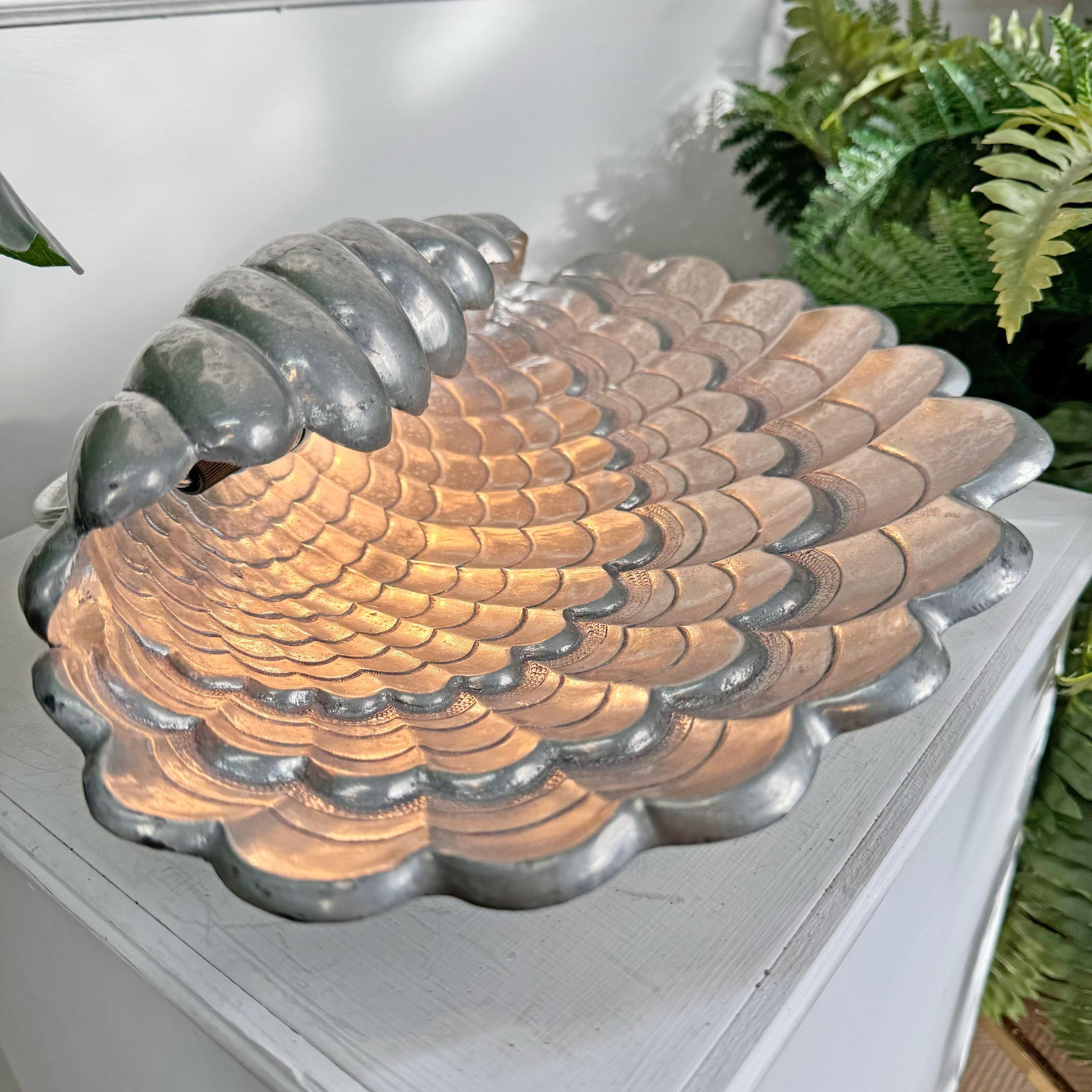 A Vincent Garnier scalloped metal shell lamp with a textured, scale-like pattern, shown resting on a white wooden surface with green plants in the background.