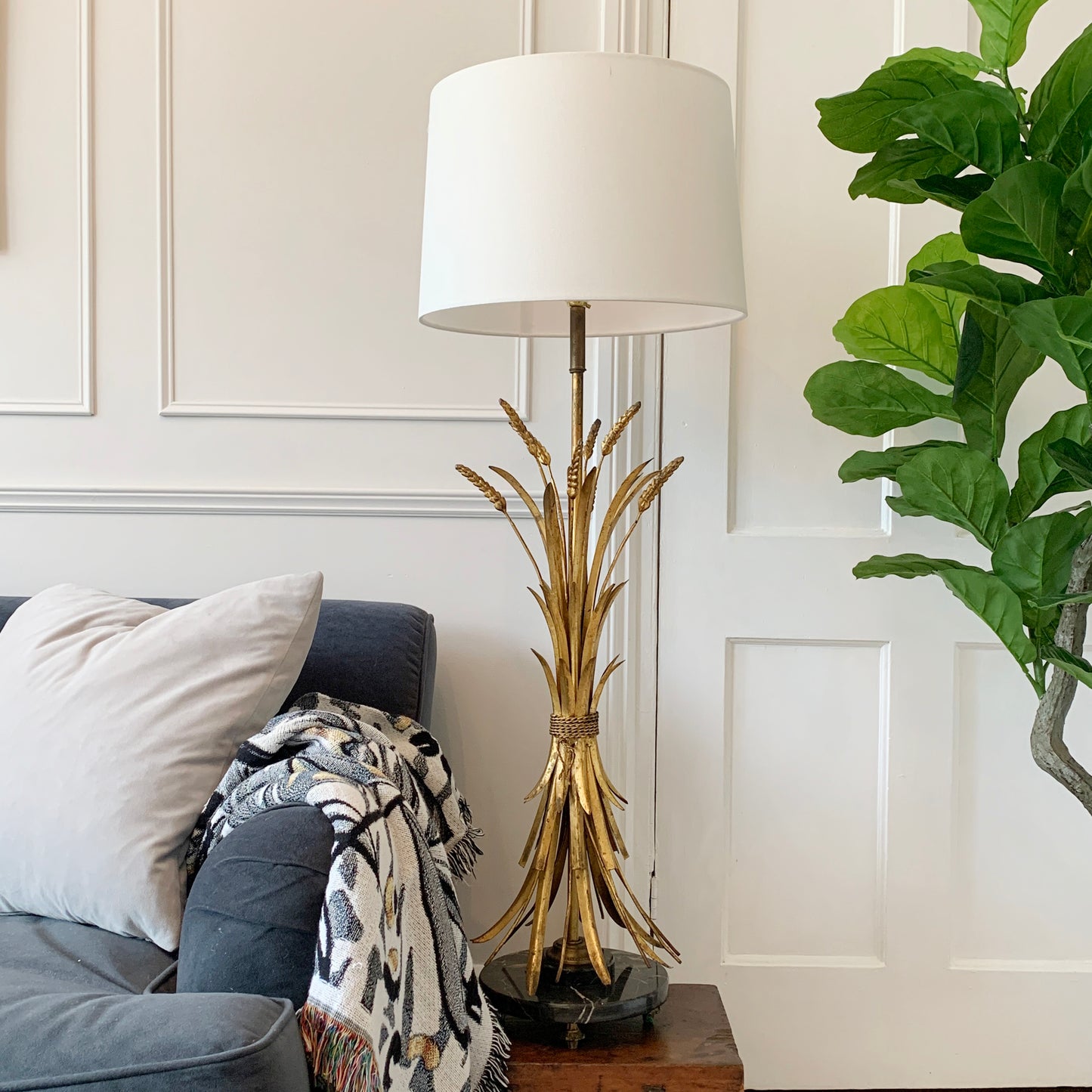 A table lamp with a white drum shade and a sculptural gold base resembling dried wheat stalks, styled on a wooden side table next to a gray sofa and a green potted plant.