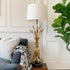 A table lamp with a white drum shade and a sculptural gold base resembling dried wheat stalks, styled on a wooden side table next to a gray sofa and a green potted plant.