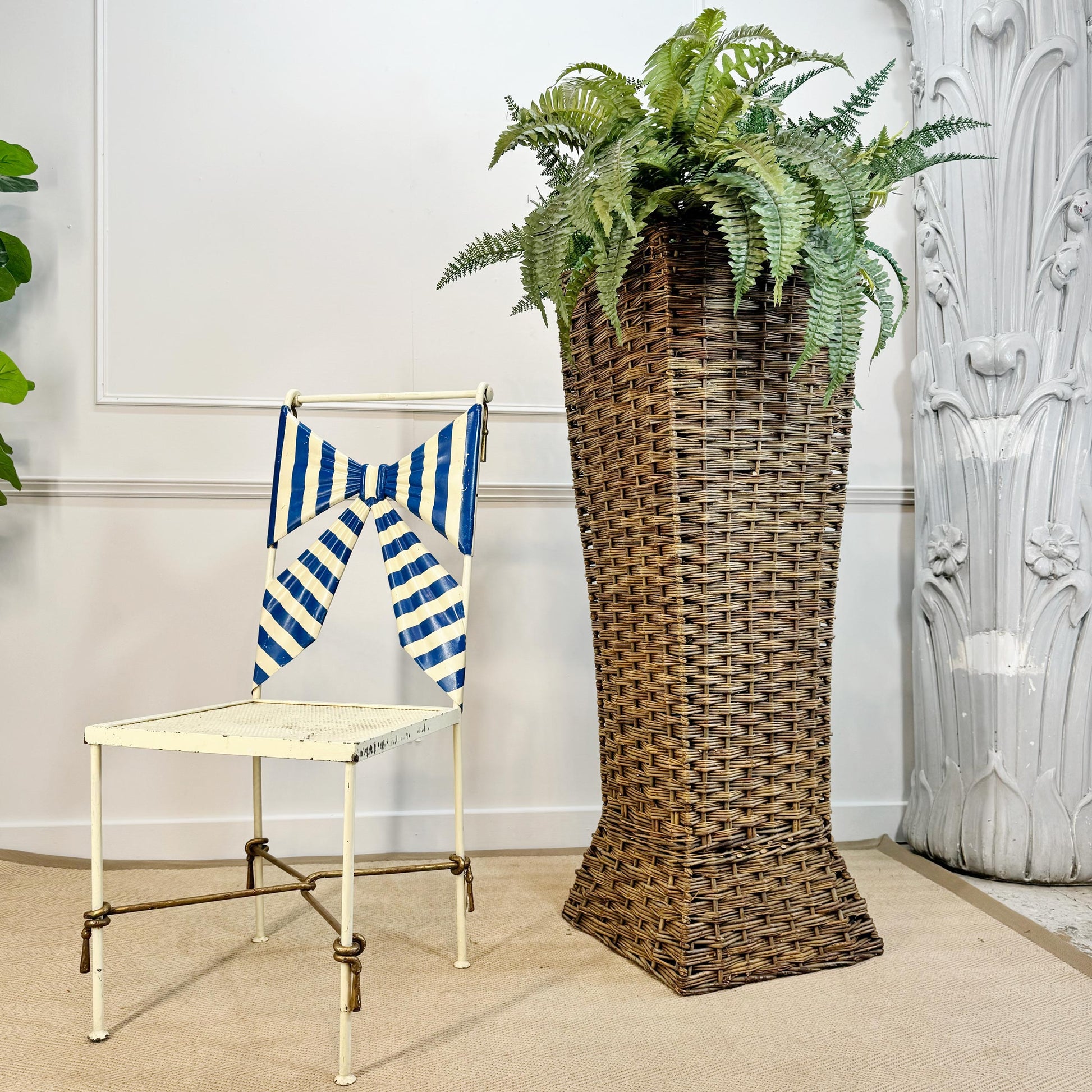 A tall, narrow brown woven planter basket holding a green fern, placed beside a white chair with a blue-and-white striped bow in an indoor room setting.