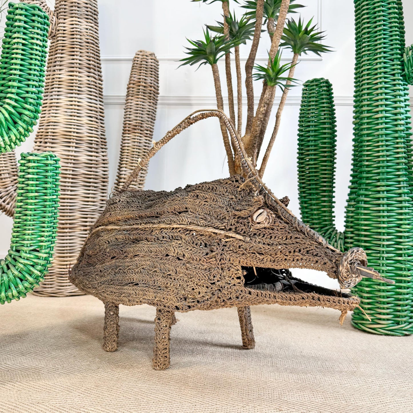 A Woven Dream Spirit Wild Pig, Papua New Guinea in brown woven grass displayed on a beige floor with decorative green and beige cactus plants in the background.