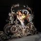An ornate gothic metal light sculpture featuring a mythical head with wings and curled scrollwork, shown lit with a warm yellow glow against a dark background.