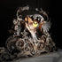 An ornate gothic metal light sculpture featuring a mythical head with wings and curled scrollwork, shown lit with a warm yellow glow against a dark background.