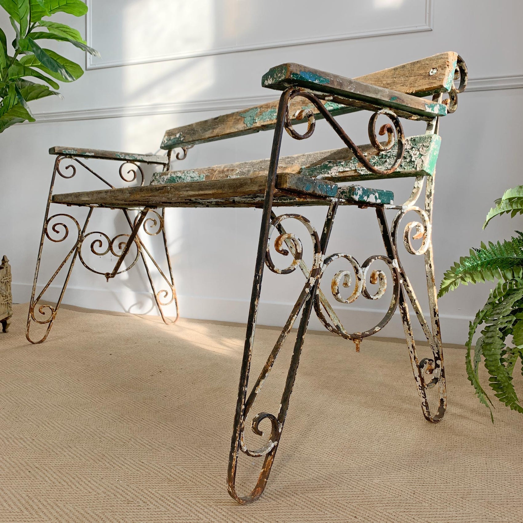 A rustic wrought iron garden bench with distressed teal and brown paint, set on a beige textured rug in a room with white paneled walls and potted green plants.