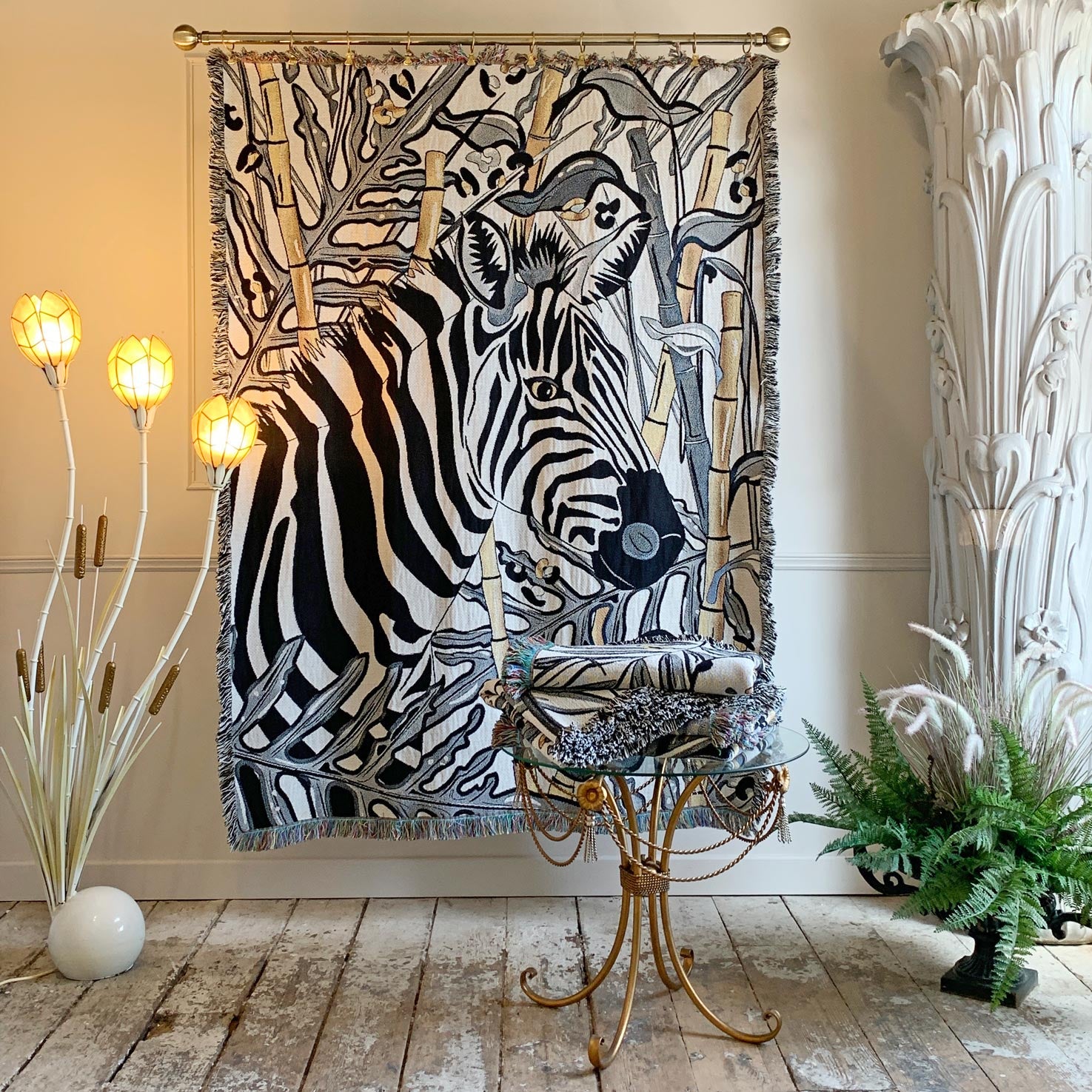A large woven tapestry featuring a detailed black and white zebra illustration with beige and gray botanical elements, hanging on a gold rod in a styled room with decorative plants, a gold side table, and warm wall lighting.