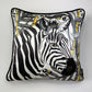 Square decorative silk pillow featuring a stylized black and white zebra print with gold accents, finished with a black piped edge, shown against a plain light gray background.